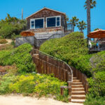 Exterior view of wooden cottage with wooden staircase from sand surrounded by natural greenery.