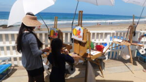 Mother and daughter on a beach deck, painting on easels underneath umbrellas