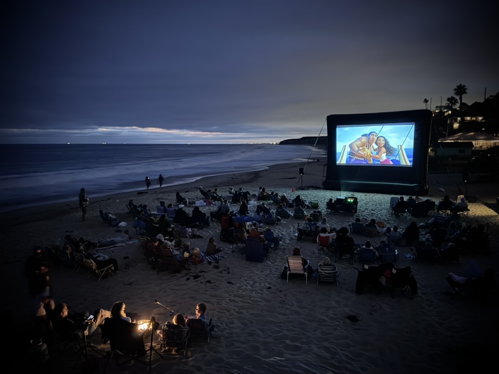 Large audience sitting on the beach with chairs and bonfires, facing a film screen at night