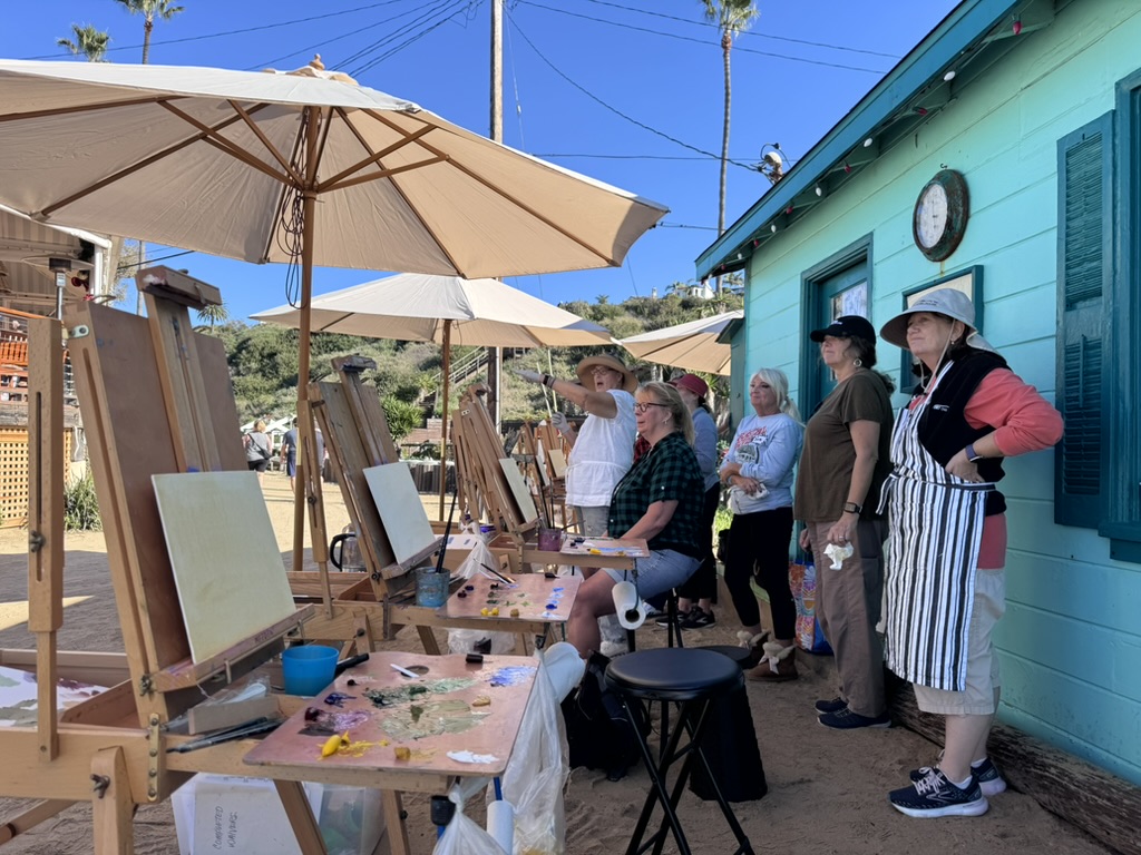 Group of people painting on easels, near umbrellas and a teal beach cottage