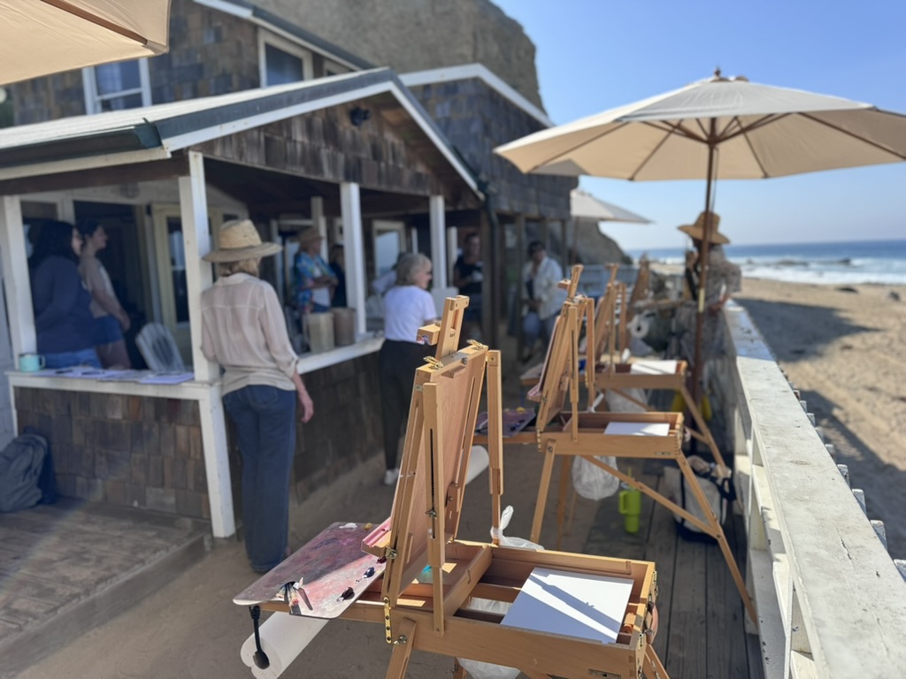 Group of people on a brown beach-cottage porch, near art easels and and umbrellas