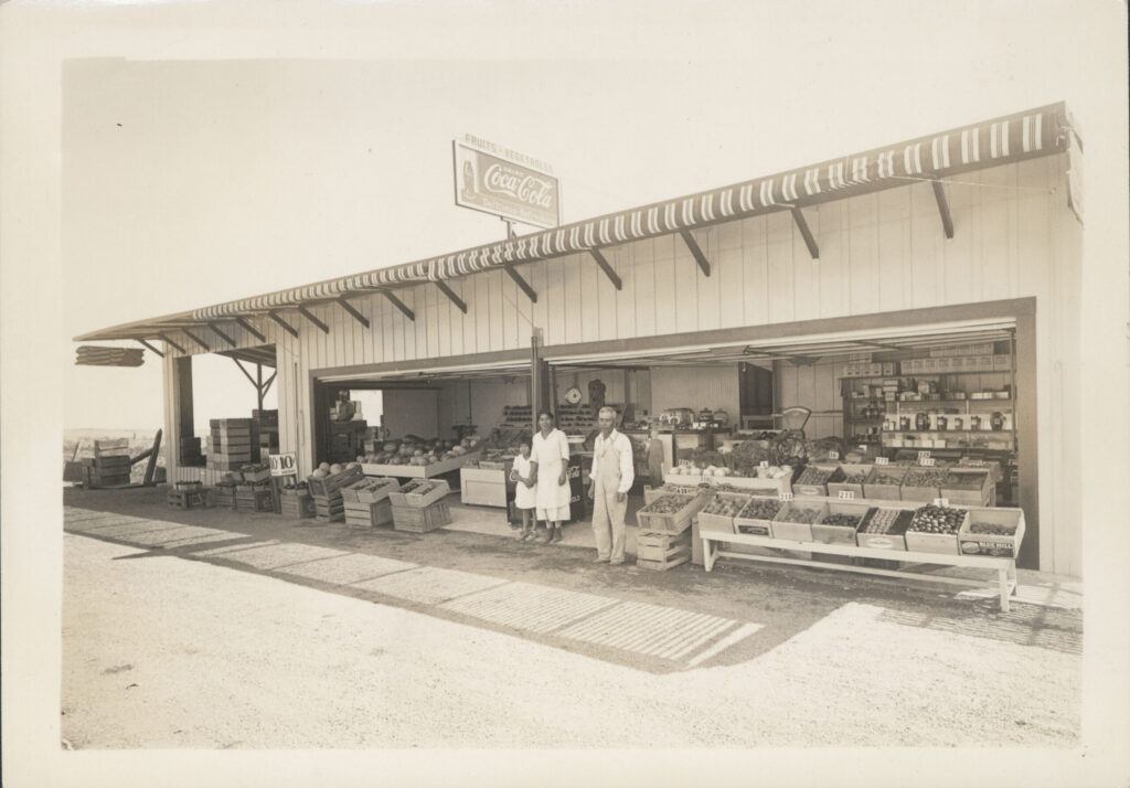 Japanese American farmers sit in front of their fruit stand in a black and white photo.