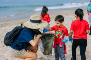 Children in red shirts and blue gloves putting beach trash into a bag carried by an educator