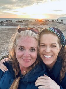 A blonde woman and red-haired woman smiling in a sunset beach selfie