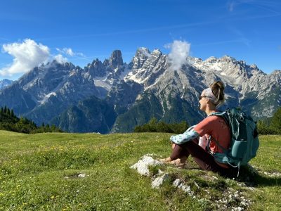 Blonde woman sitting on a field in front of large mountains, wearing a backpack